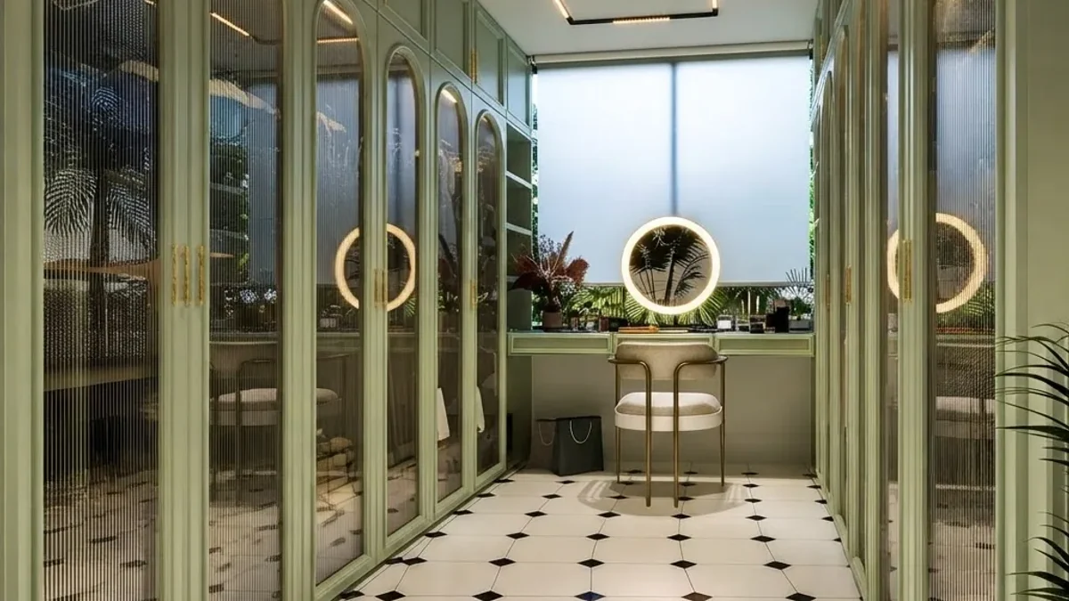 Sage green walk-in closet with arched reeded glass wardrobe doors and gold round pulls, a vanity under the window with a round lit mirror and a black and white tiled floor