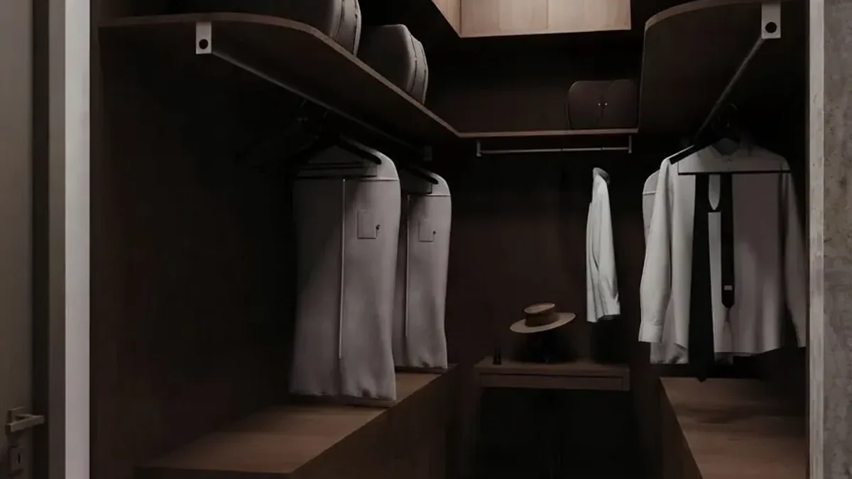 Compact dark walnut walk-in closet with a lit hanging rail holding garment bags and white shirts, a shelf above with a hat, and drawers below