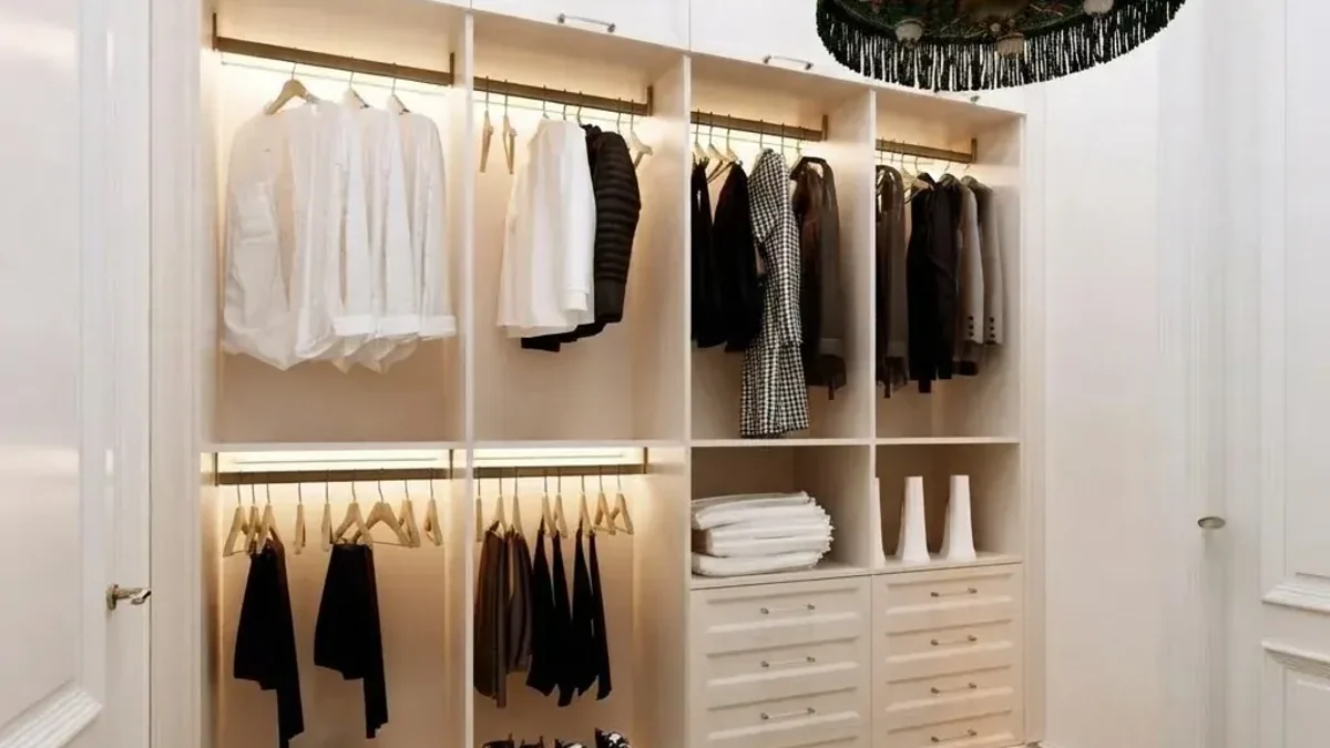 Traditional white walk-in closet with paneled walls, open cream joinery with double hang rails lit from above, folded stacks on shelves and a column of white shaker drawers