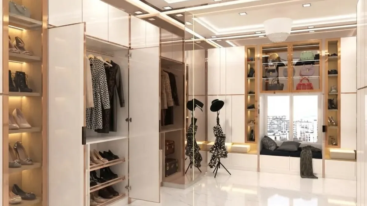 Bright white walk-in closet with lit wood-lined shoe shelves floor to ceiling on the left, open hanging behind a glass door and a cushioned window seat with drawers under it at the back