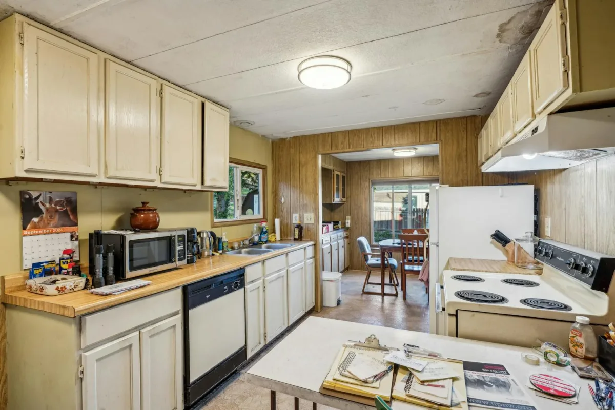 Before photo of a dated galley kitchen with cream cabinets, laminate counters, wood panelled walls, a coil-top range, white fridge and a folding table, looking through to a small dining nook