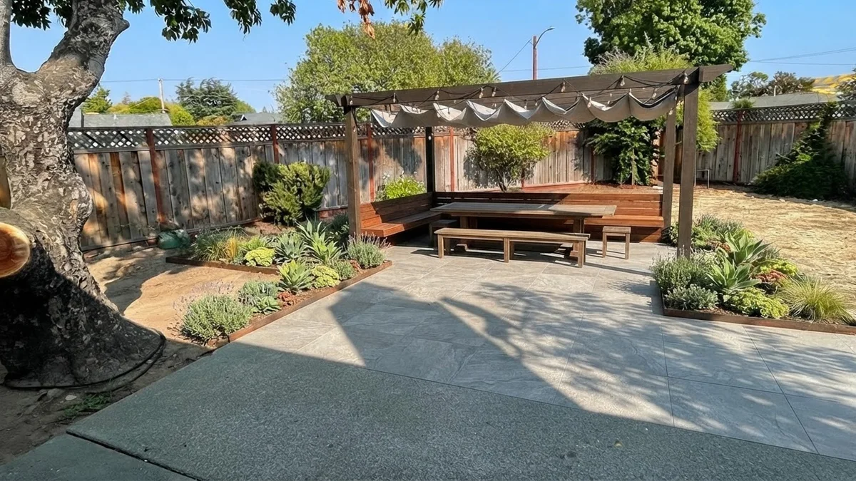 The base backyard rendered with an extended porcelain paver patio, a timber pergola, a long dining table and festoon lights