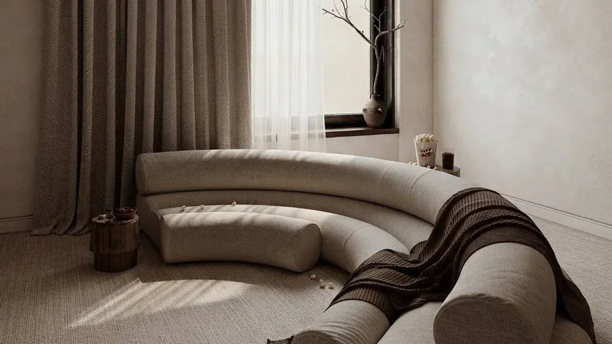 Cream curved sofa with a long bolster cushion and a dark striped throw in a plaster walled room, with floor to ceiling taupe curtains, a deep window reveal holding a bare branch in a vase and hard sunlight raking across a textured beige rug