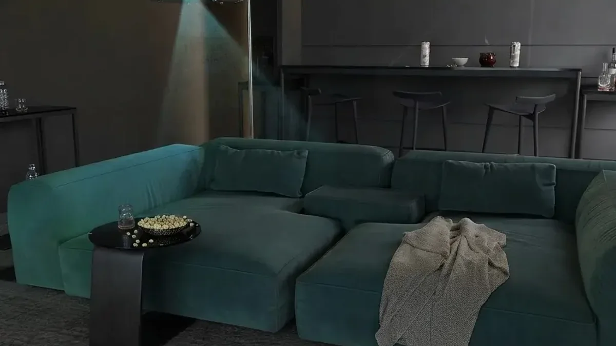 A dark lounge with a deep teal modular sofa and a knitted throw, a ceiling mounted projector casting a visible beam, a small black side table holding a bowl, and a bar counter with stools in the background