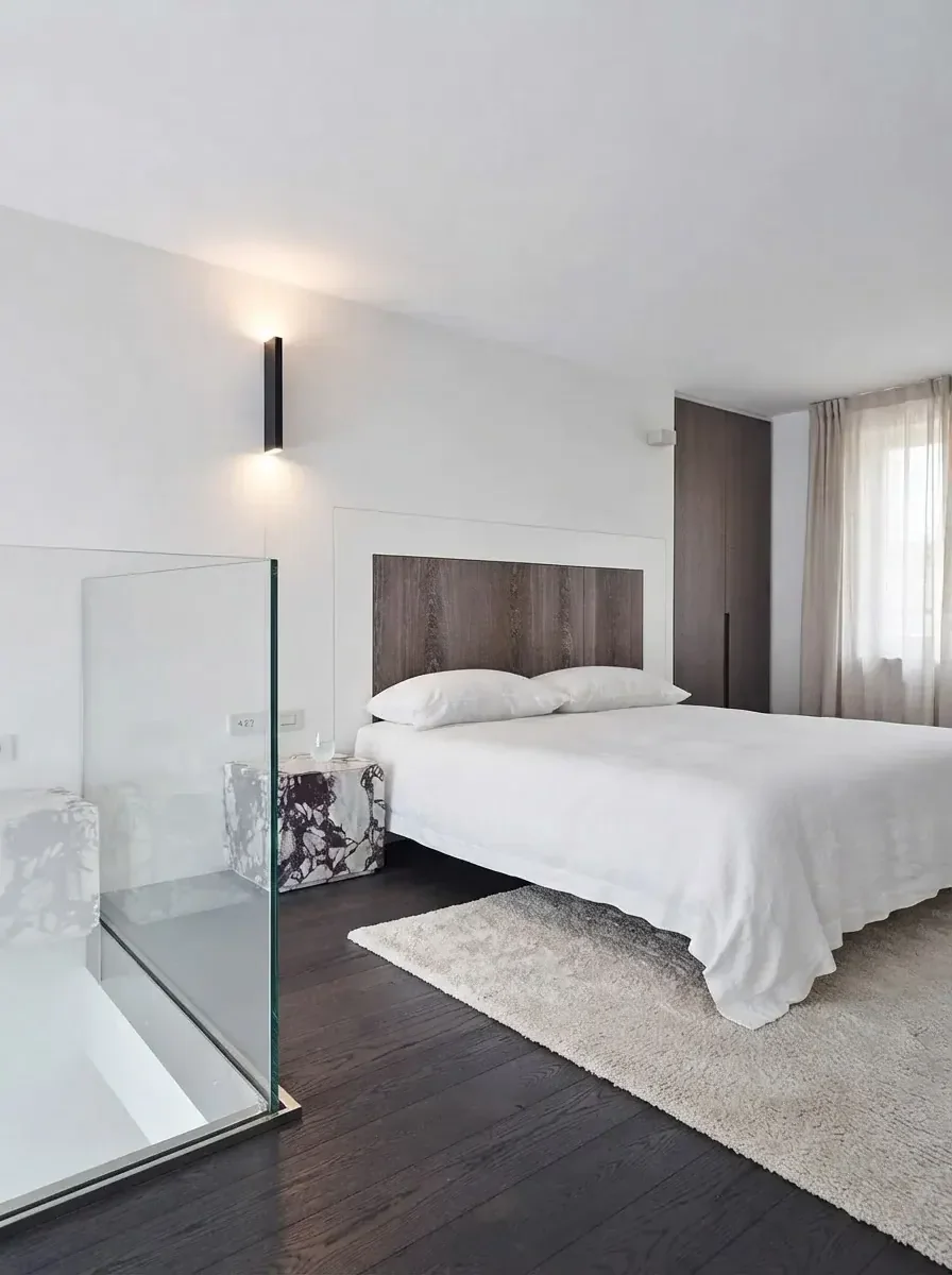 Compact white bedroom render with a double bed pushed against the right wall under a dark oak headboard, a glass partition on the left and a rug at the foot