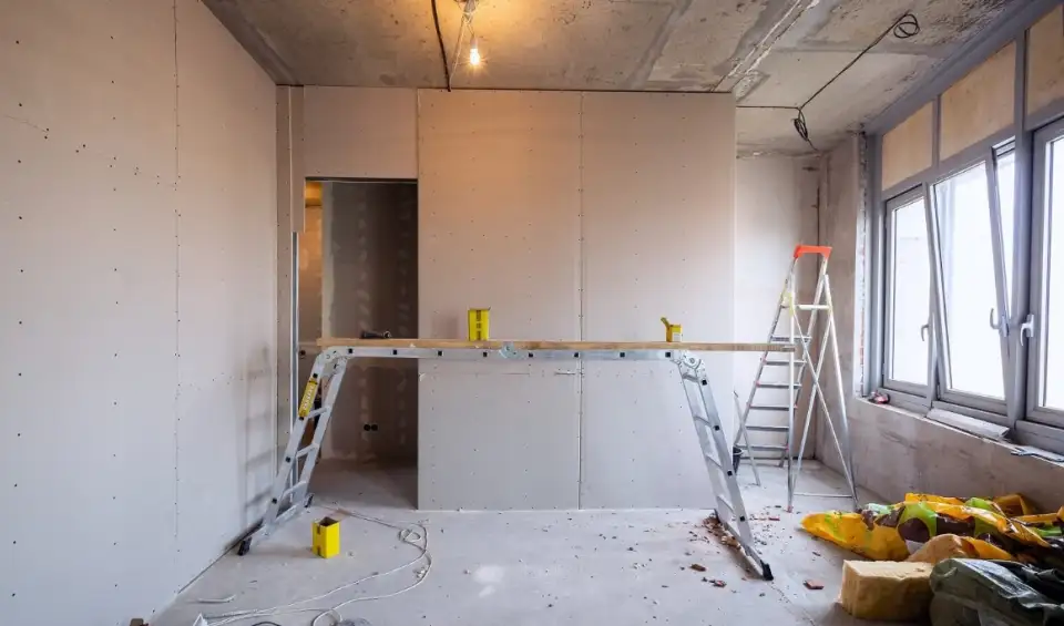 Construction room with raw drywall, ladders, concrete floor, and exposed ceiling before renovation