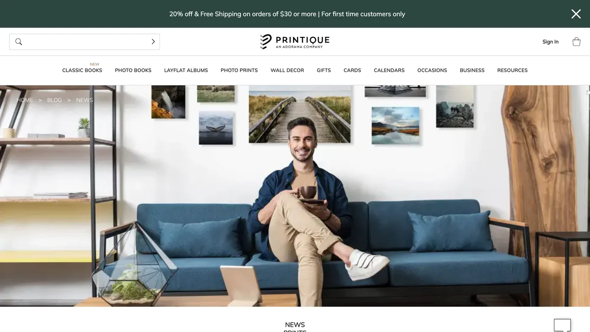 Printique blog page titled Introducing The Gallery Wall Design Wizard showing a man seated on a blue sofa beneath a gallery wall of framed photographs