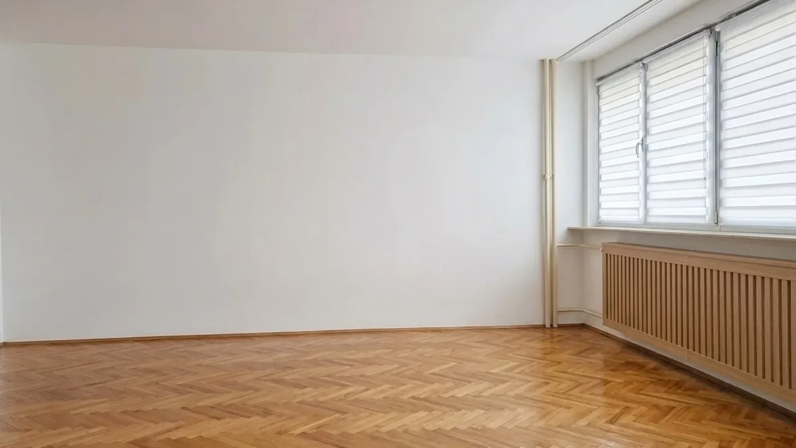 Before renovation: real empty room in an older apartment with herringbone parquet, exposed pipes, old wooden radiator cover, and dated blinds