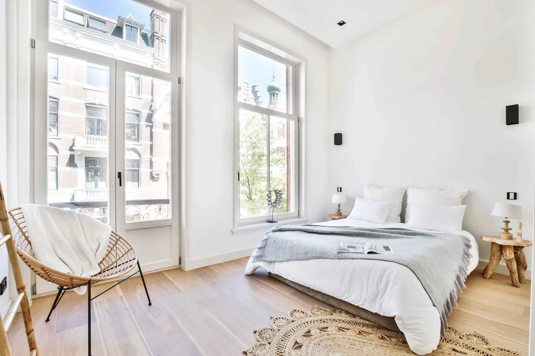 Original bedroom with white bedding, rattan accent chair, jute rug, and wooden side tables — the reference before AI redesign