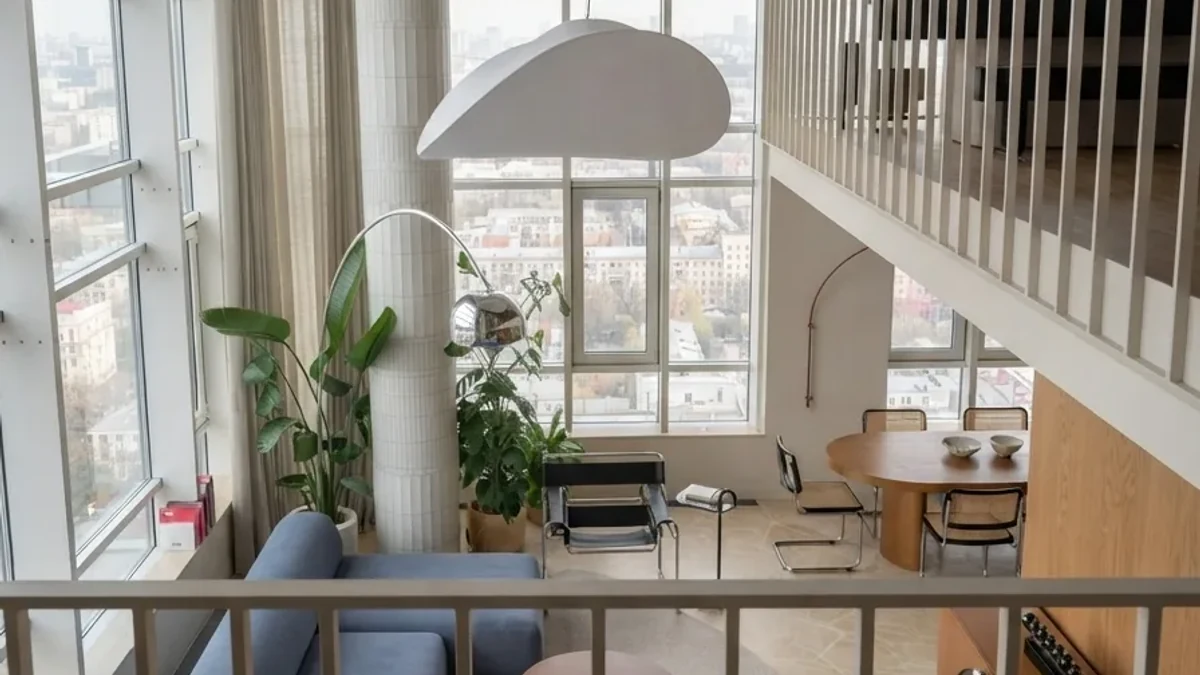 Loft living room design idea: a double-height apartment with floor-to-ceiling city windows, a blue sofa, cantilever dining chairs, large potted plants and a sculptural white pendant lamp