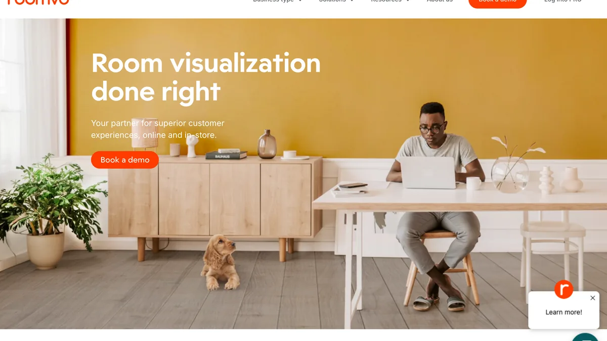 The Roomvo website headlined Room visualization done right, with a Book a demo button, a man working at a laptop in a room with a mustard wall and grey wood-look flooring, and a puppy on the floor