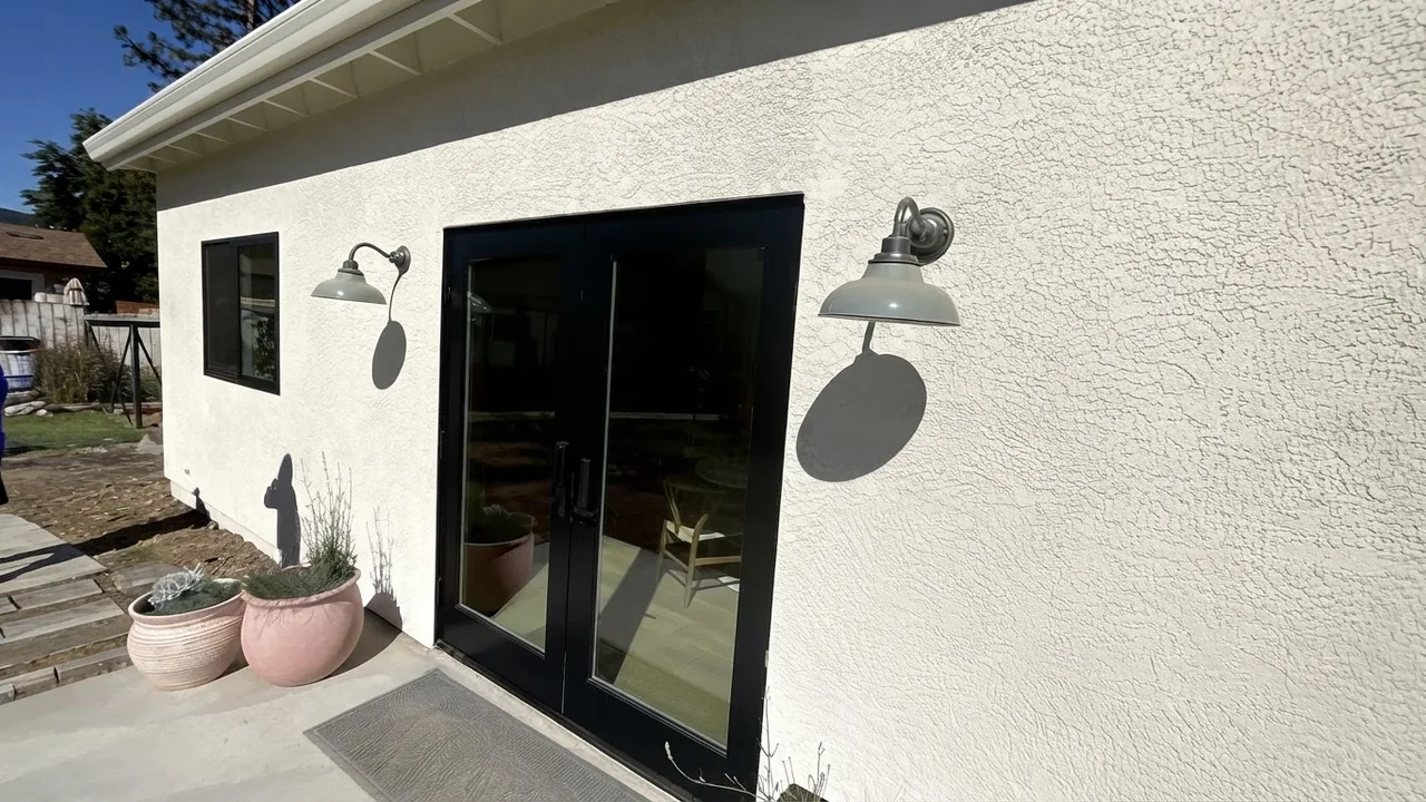 The finished entrance of a converted garage unit: white stucco wall, black framed glazed double doors, two black gooseneck wall lights and potted plants on a paved threshold