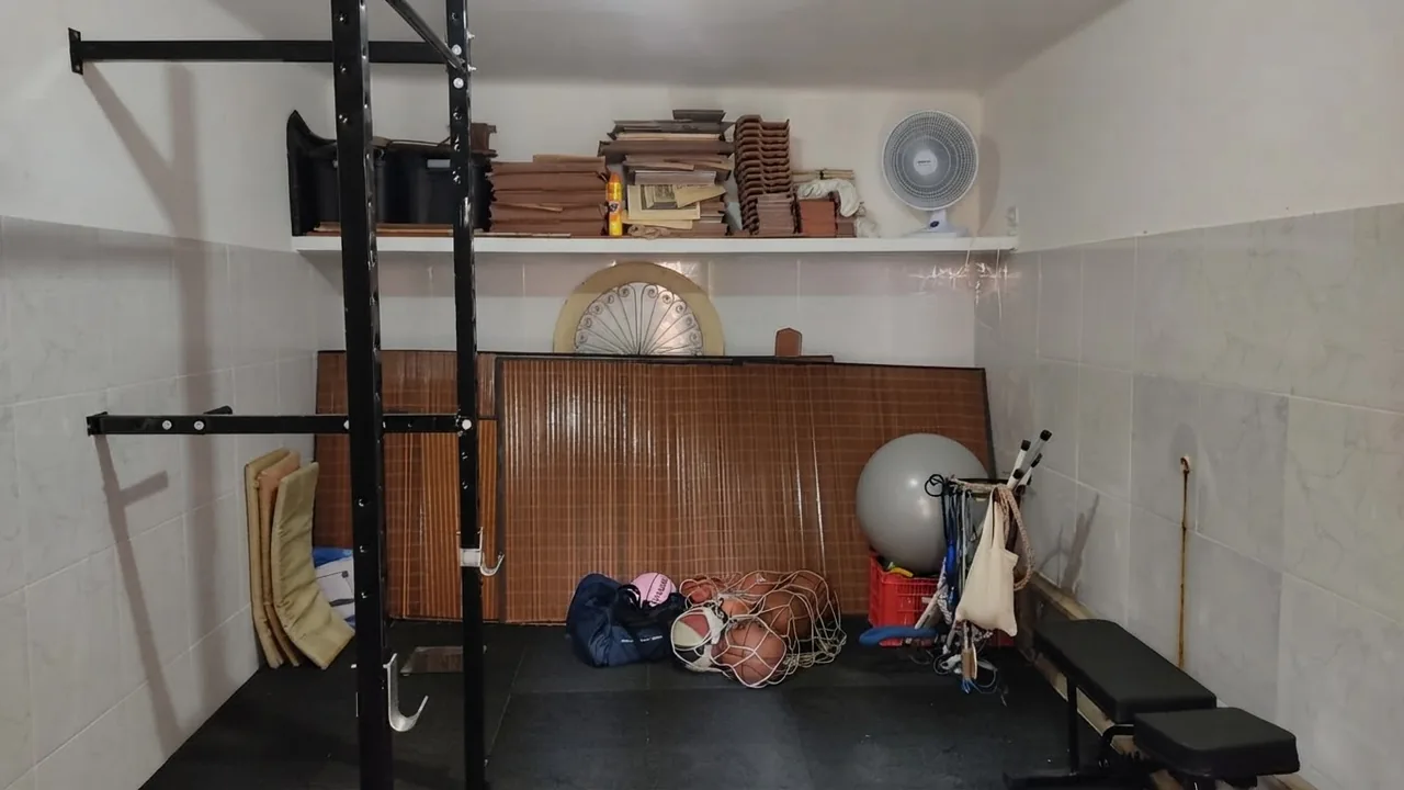A real garage before conversion, with wall shelving full of boxes and books, a bicycle wheel, an exercise ball, bags on the floor, a bamboo screen and a rack against bare walls
