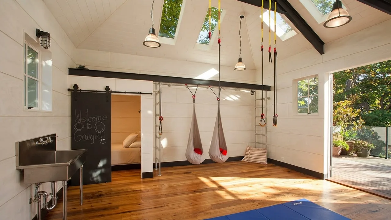 Inside the same converted garage: white walls, a sprung timber floor, skylights, an exposed dark steel beam, gymnastic rings, a barn door, a utility sink and a climbing ladder