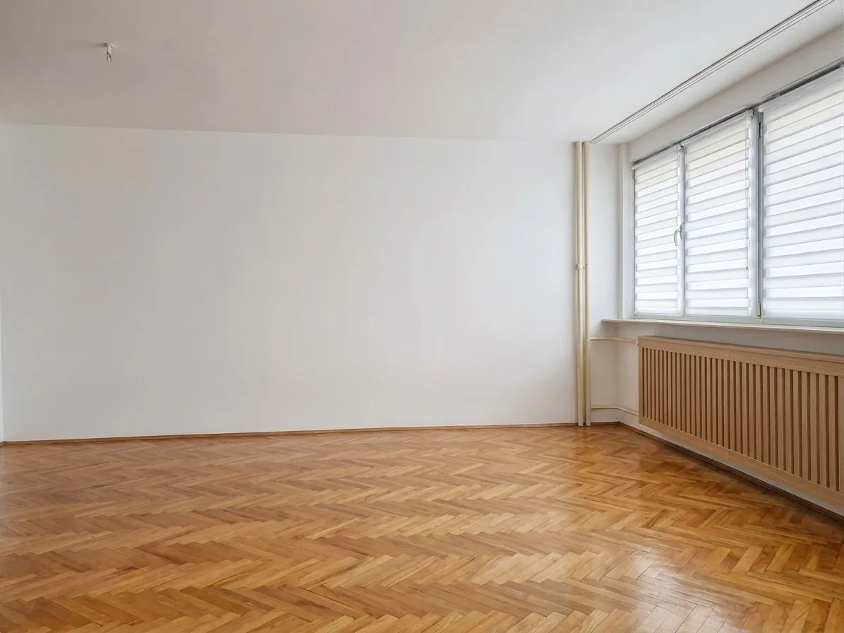 Empty living room with herringbone parquet floors, white walls, and radiator under window — typical house flip property before renovation and staging