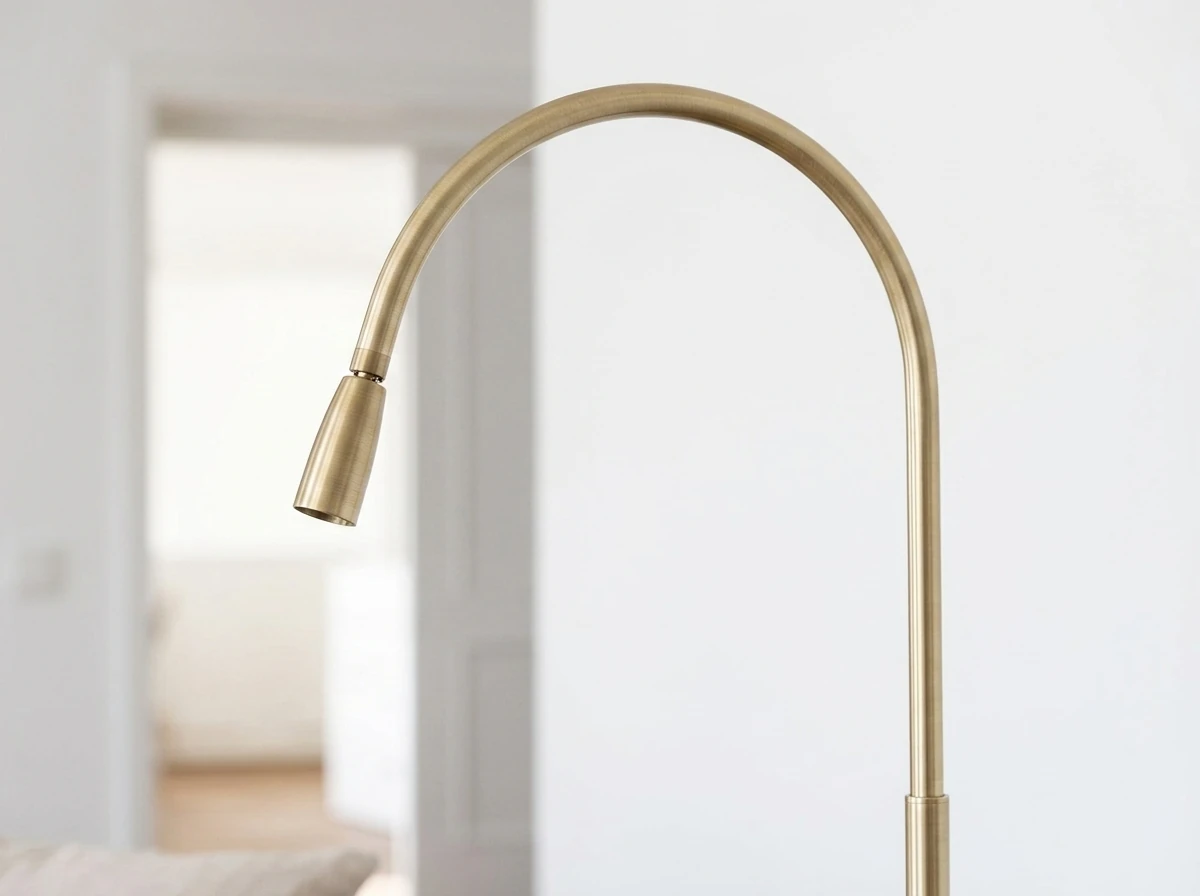 Close-up of the head of a slim brushed-brass arc floor lamp against a clean white wall, showing the soft satin sheen of the brushed metal and the smooth curve of the arc
