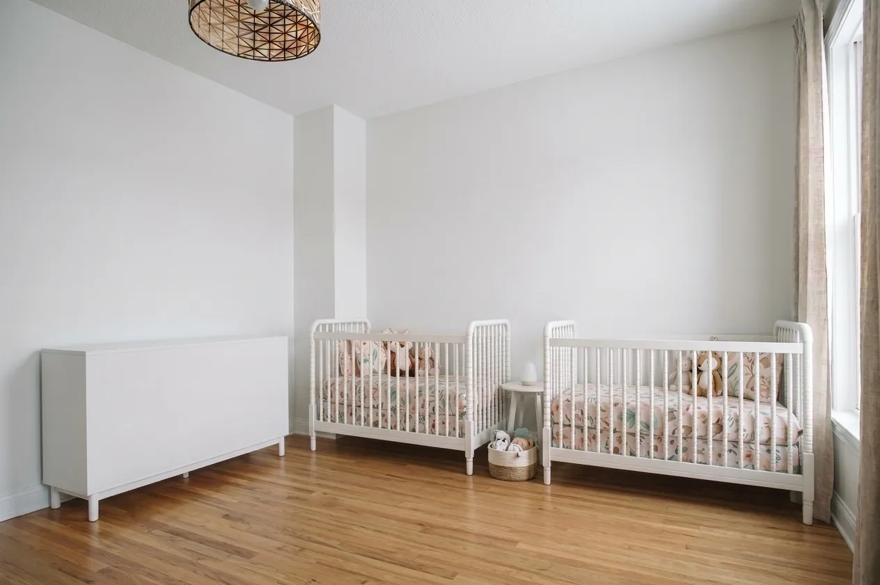 Simple twin nursery with two white cribs, white dresser, rattan pendant light, linen curtains, and minimal decor on light hardwood floors