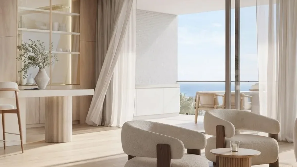 A pale limewashed living room with a fluted plaster console table, sheer floor to ceiling curtains, two cream boucle armchairs around a small round table, pale timber flooring and a sea view through the glazing