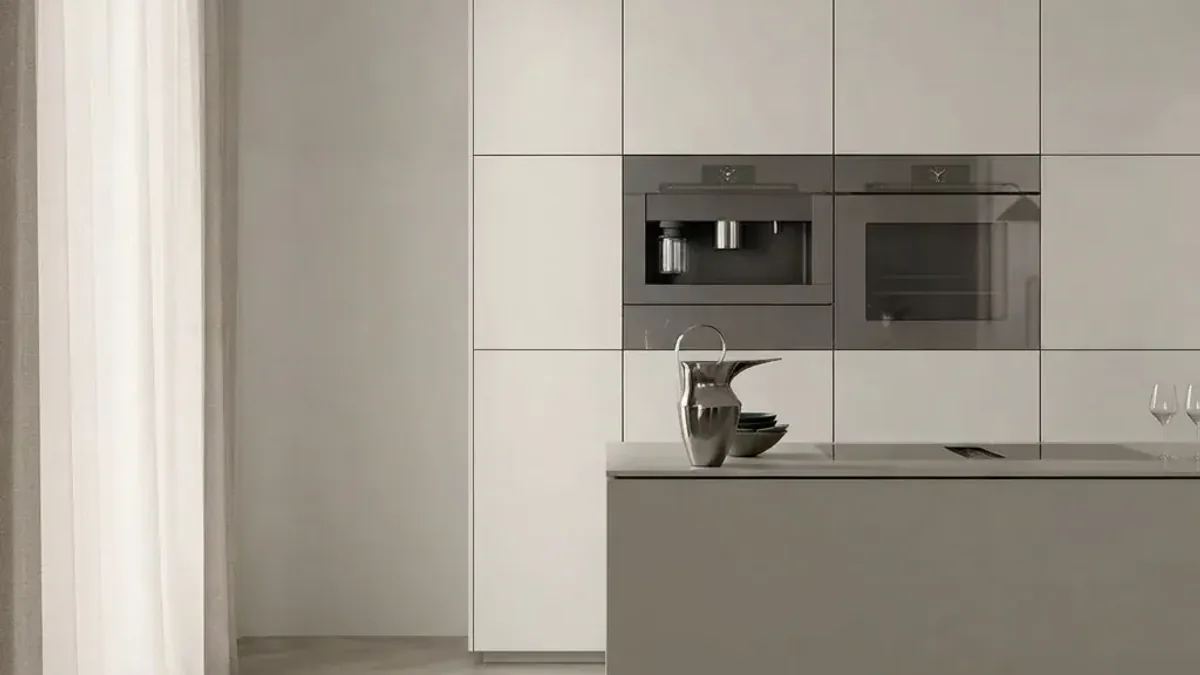 A pale minimal kitchen with a tall run of handleless cabinets holding an integrated coffee machine and oven, a low island with a flush hob, a stone jug and bowl on the worktop and a sheer curtain at the window