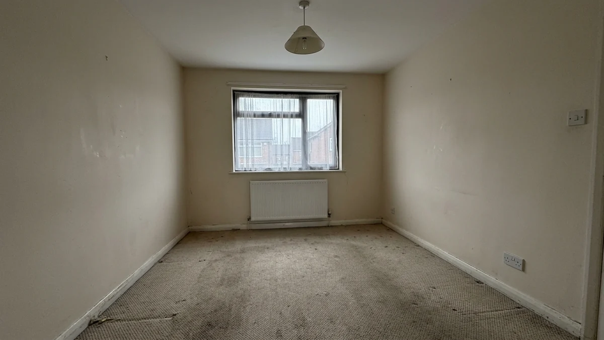 A typical unstaged estate agent photograph of an empty living room under flat grey light with worn carpet and blank walls
