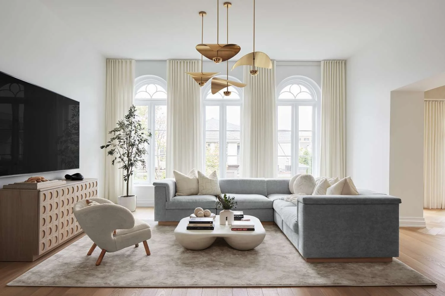 Original photo of a character home living room with three tall arched windows, light gray sectional sofa, sculptural gold chandelier, white cloud coffee table, and cream armchair