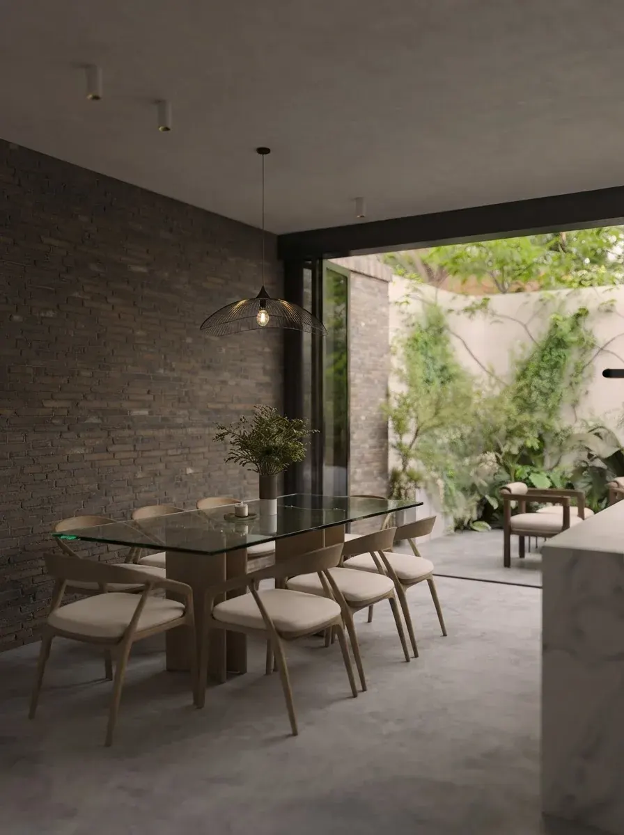 Garden room with exposed brick walls, a glass table with woven chairs, a wide glazed opening onto a planted courtyard and a pendant light overhead