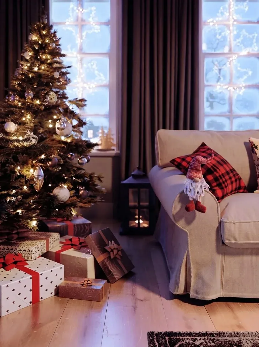 Close view of the same cottage tree with warm lights, a lantern and red and white wrapped gifts beneath it beside a white slipcovered sofa