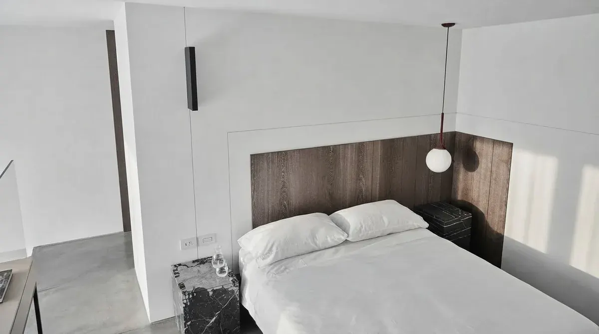 Modern minimalist bedroom interior design: white bedding on a simple bed, a dark oak panel as the only headboard, a marble cube nightstand and a single globe pendant against bare white walls