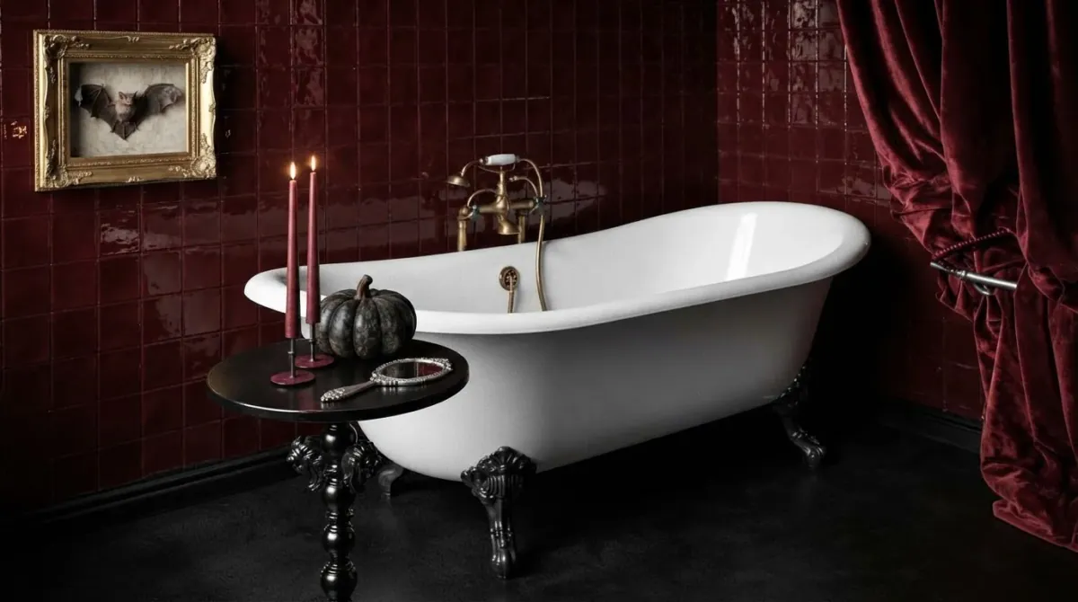 Gothic Halloween bathroom with glossy oxblood tile, a white clawfoot tub with black feet, a dark red velvet curtain, two red taper candles and a black pumpkin on a turned black side table, and a gold framed bat print on the wall