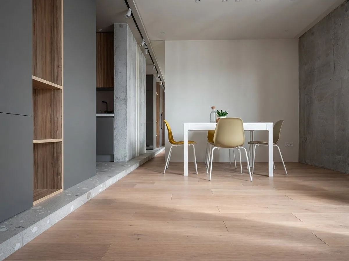 Small apartment where a seamless grey microcement kitchen floor meets a light oak floor under a white dining table, with oak and grey cabinets and a plain oak door
