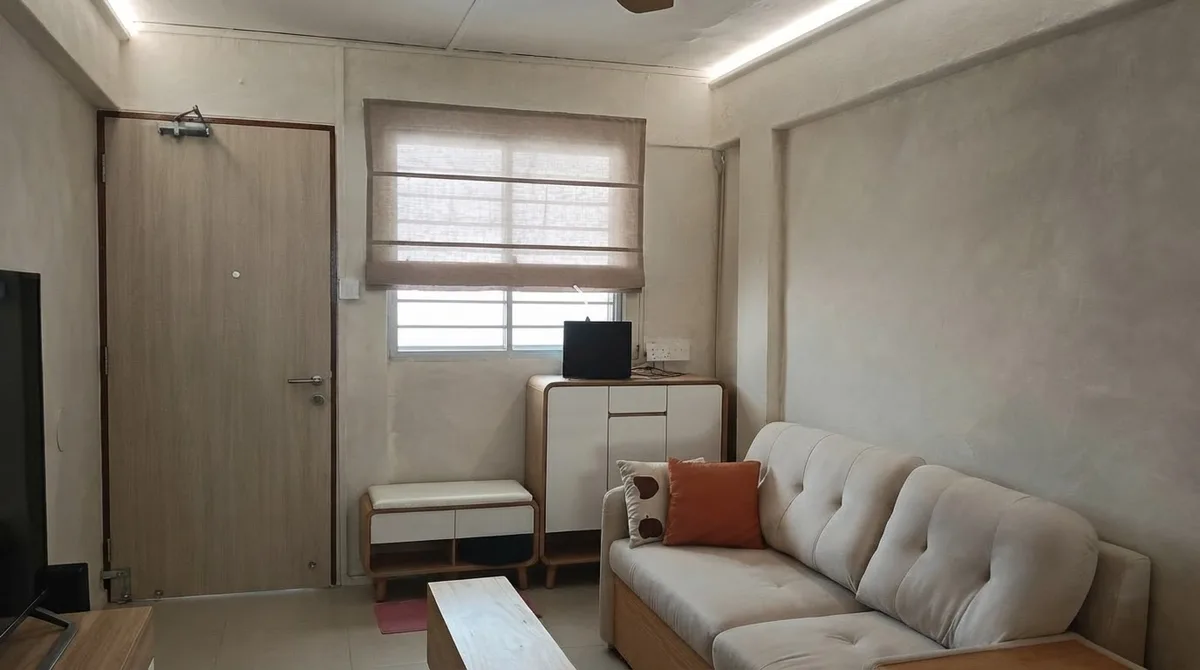 A small apartment living room with pale microcement walls, a cream tufted sofa against the right wall, an orange cushion, a wood cabinet under the window and a plain oak interior door on the left