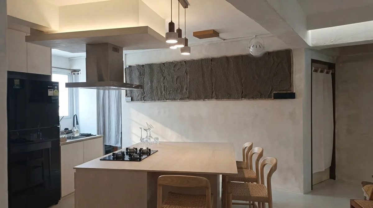 Pale microcement kitchen and dining area with a light oak island, black cooktop, rattan chairs and a white ceiling fan above the dining table