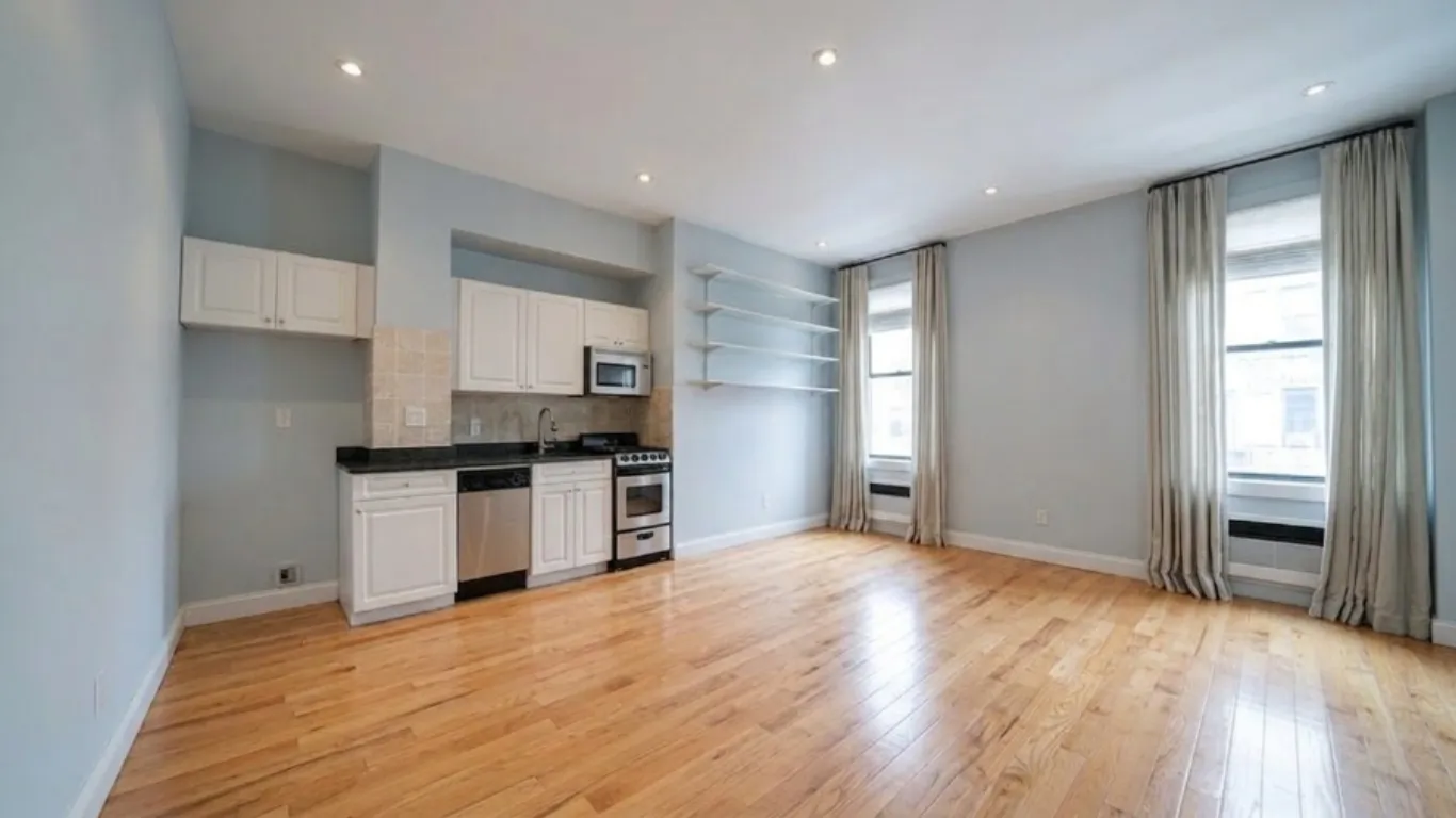 Empty studio apartment with kitchenette, hardwood floors, blue-grey walls, and two large windows before furnishing