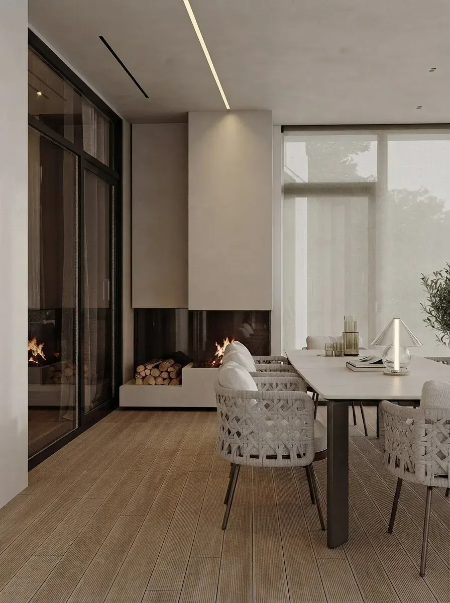 Glazed sunroom with a built in fireplace, a pale dining table with woven chairs, full height glass on two sides and fog over the garden beyond
