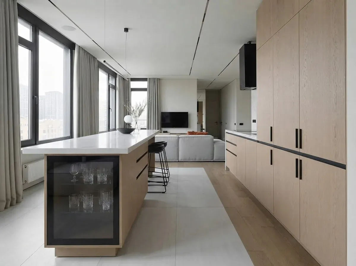 A long pale oak kitchen with tall cabinets and black bar handles along one wall, a white-topped island with a wine fridge facing it, big windows and the living room beyond