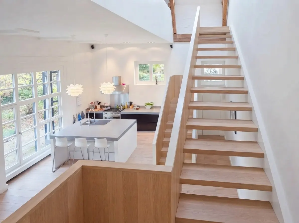 Open timber staircase rising to a barndominium loft with a plain white guard, a pale kitchen with an island and bar stools below, under a vaulted ceiling