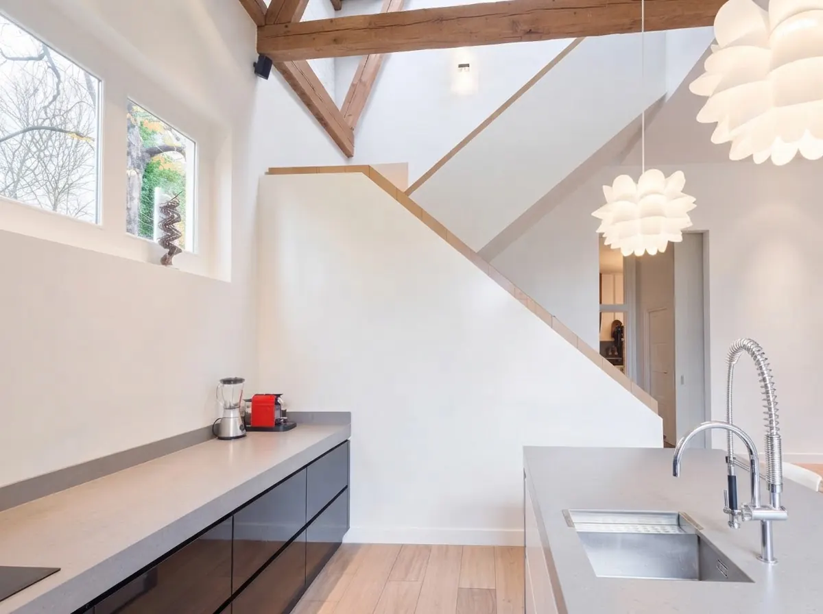 Bright barndominium kitchen under a white vaulted ceiling with exposed timber beams, a pale island with bar stools and a window overlooking trees